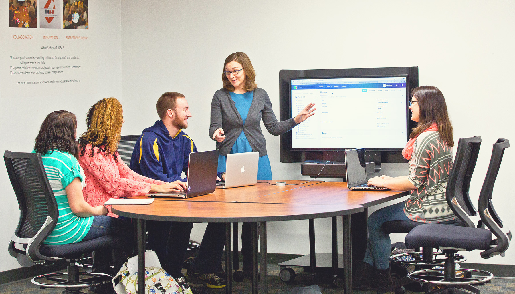 a professor and four students around a table with a smart board.