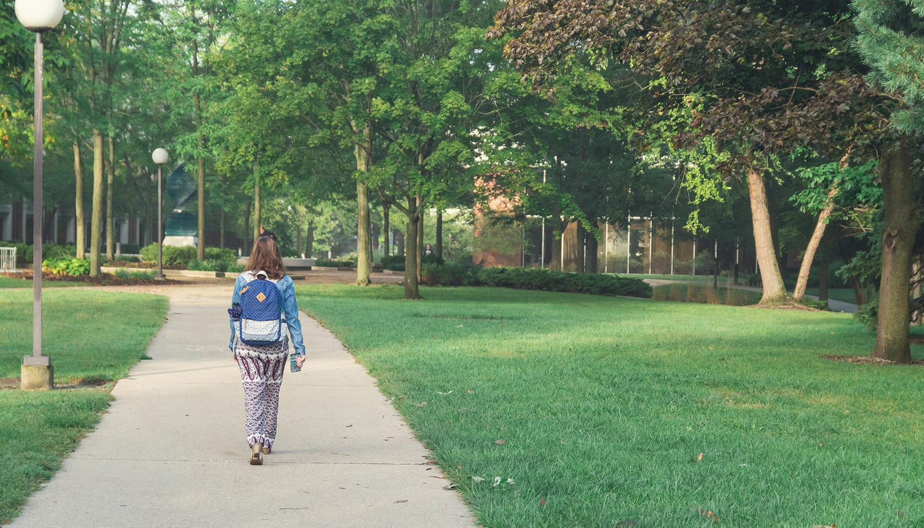 Female student walking the sidewalk among green grass and trees.