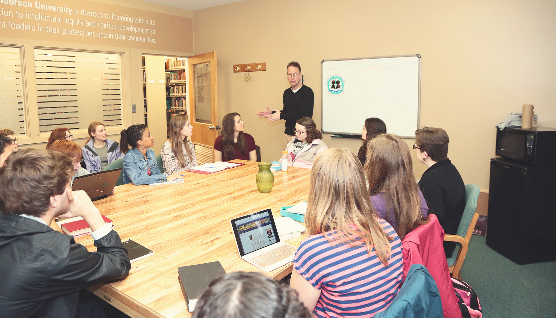 a male professor talks to a class seated around a large wooden table