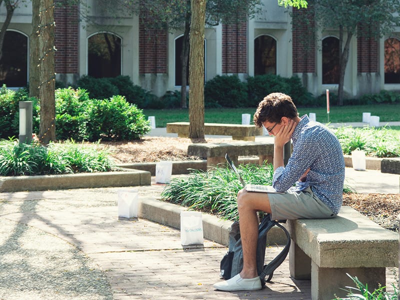 male student with laptop sitting on a bench outside