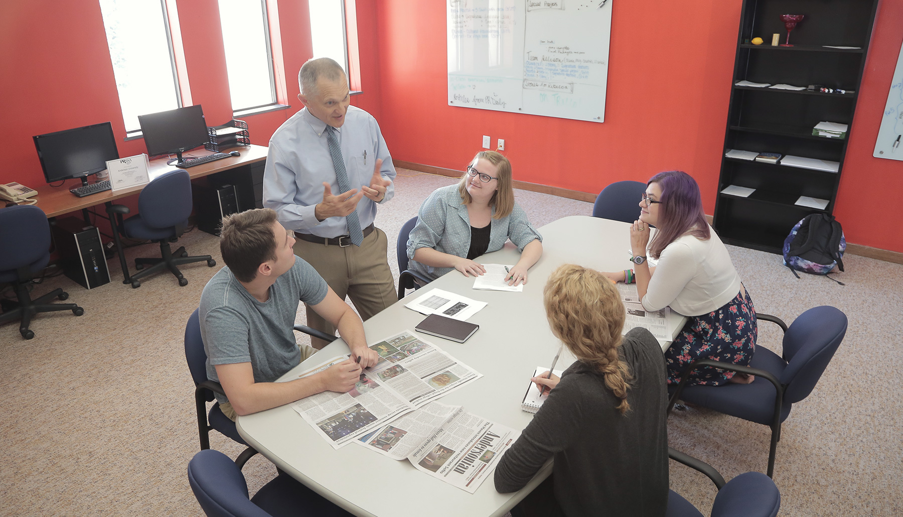 a male professor talks with four students - three female, one male - around a table with newspapers on it