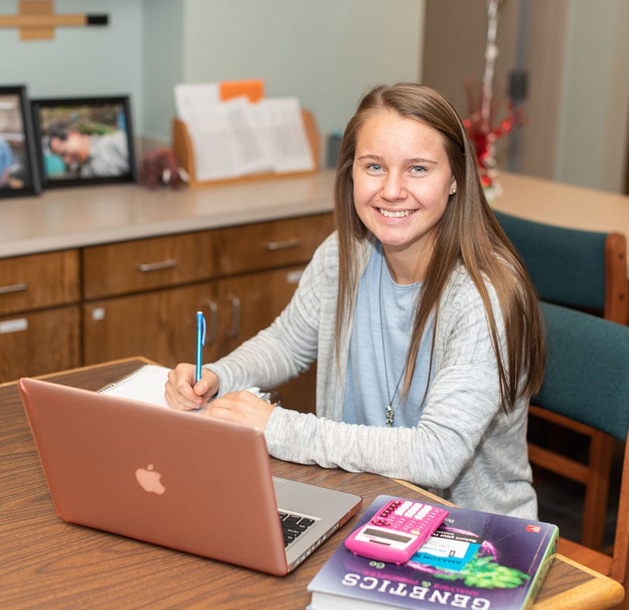 a young woman with blue eyes sits at a table with a Macbook, a genetics textbook, and a calculator.