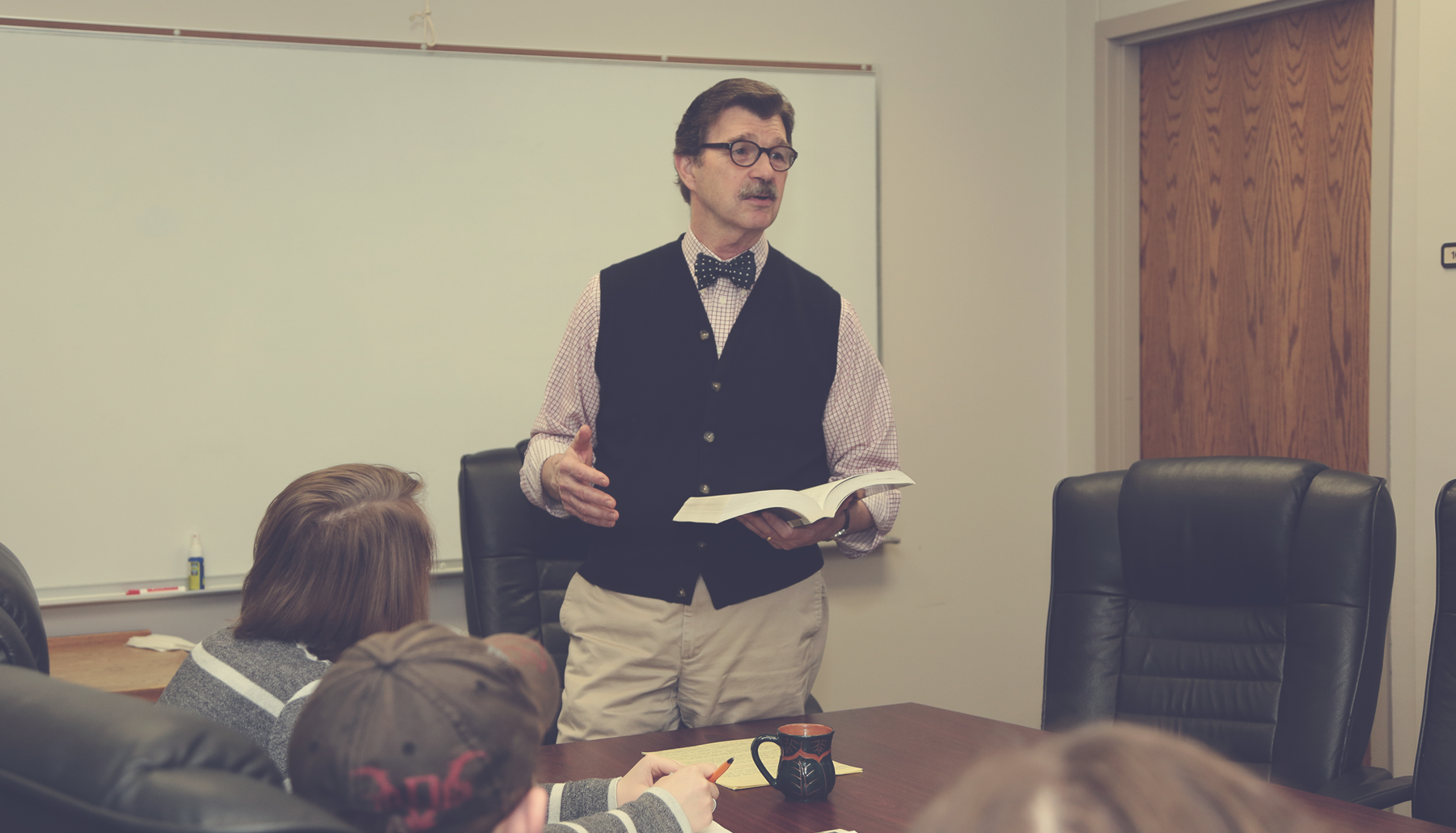 a professor wearing a bowtie speaks to a group of students seated at a table